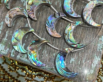 May include: Abalone shell crescent moon pendants on a wooden surface. The iridescent pendants are strung on thin silver-colored wire. The moons display a range of colors, including blues, greens, and purples, with a textured surface.