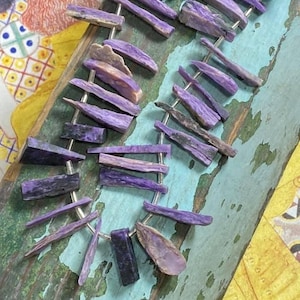 May include: A strand of purple and silver gemstone beads. The beads are long and thin, with a slightly iridescent sheen. They are strung on a silver wire.