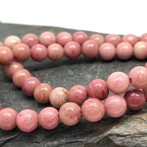 Puede incluir: Una hebra de cuentas de piedras preciosas rosadas, probablemente de rodonita, dispuestas en un patrón circular. Las cuentas son lisas y pulidas, con un brillo sutil.