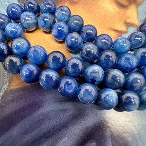 May include: A necklace of polished blue kyanite beads. The beads are spherical and display a marbled effect with different tones of blue. The background has a soft, artistic image with a celestial theme.