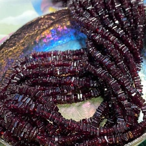May include: A strand of dark red gemstone beads, each bead is a rectangular prism shape. The beads are piled in a bowl.