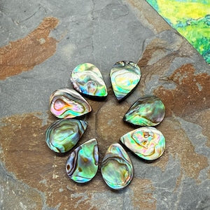 May include: Eight iridescent abalone teardrop-shaped pendants arranged in a circle. The pendants display a mix of green, blue, and purple hues with a pearlescent sheen. They are set on a dark gray stone surface.