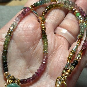 May include: A multi-colored gemstone necklace with a green beetle pendant. The necklace features a variety of beads, including pink, green, and gold. The beetle pendant is made of green stone and has gold accents.