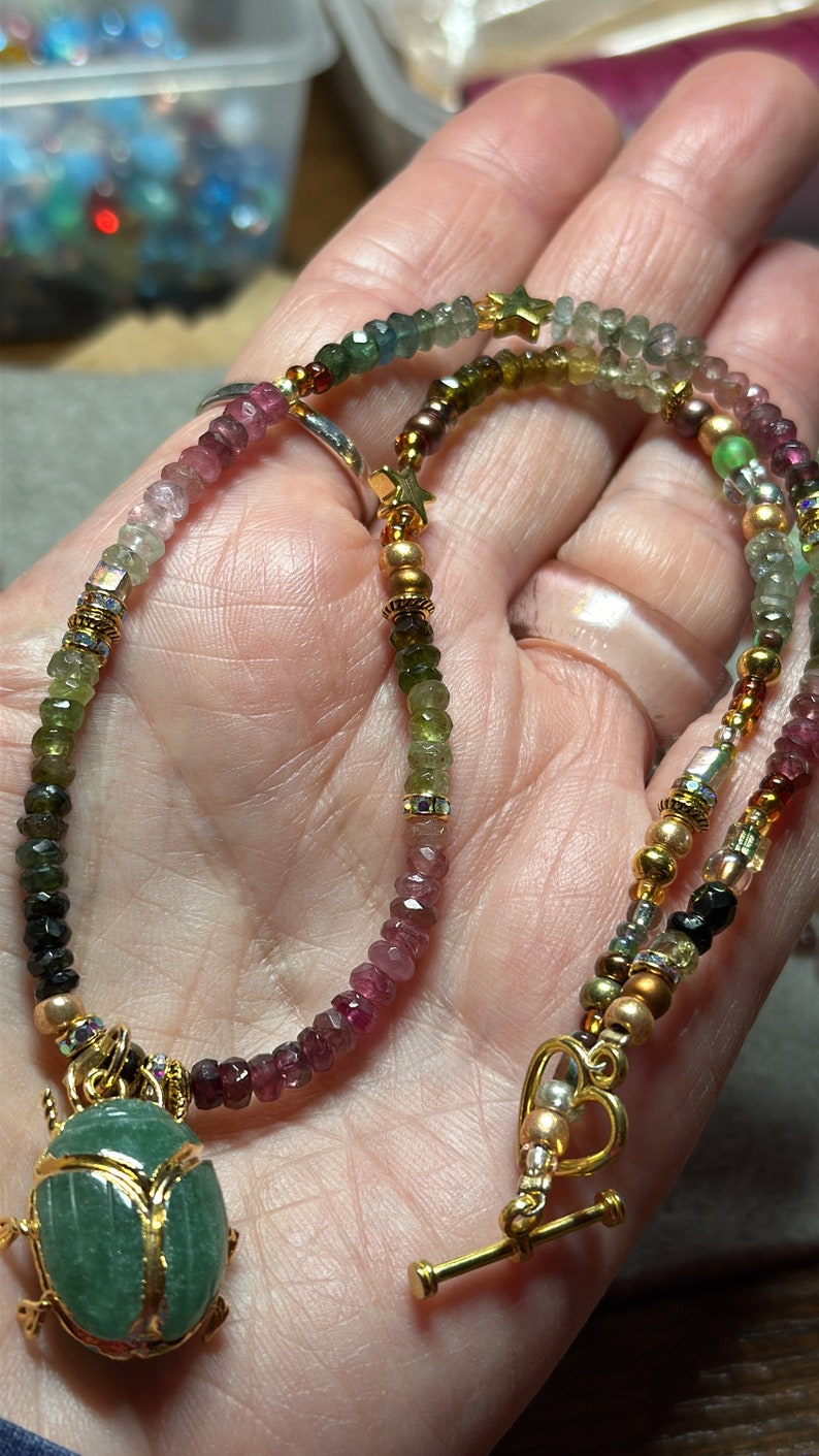 May include: A multi-colored gemstone necklace with a green beetle pendant. The necklace features a variety of beads, including pink, green, and gold. The beetle pendant is made of green stone and has gold accents.