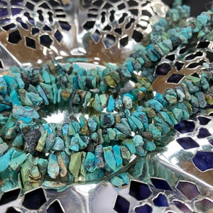 May include: A strand of turquoise chip beads, with a mix of blue, green, and brown tones. The beads are irregular in shape and size, and are arranged in a loose, flowing pattern. The beads are displayed on a silver background.
