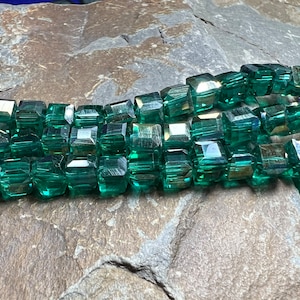 Könnte beinhalten: Eine Strähne dunkelgrüner, facettierter Glasperlen. Die Perlen sind quadratisch und haben eine glänzende, reflektierende Oberfläche.