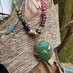 May include: A green gemstone scarab beetle pendant on a multi-colored beaded necklace. The necklace features pink, green, and black beads.
