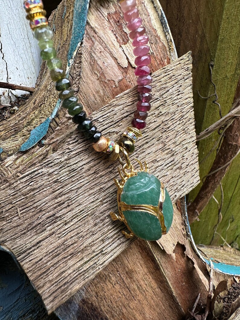 May include: A green gemstone scarab beetle pendant on a multi-colored beaded necklace. The necklace features pink, green, and black beads.