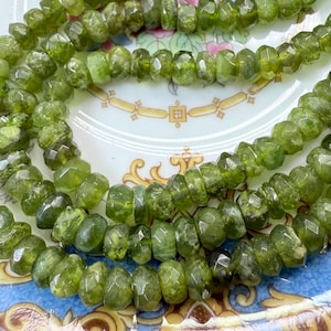 May include: A strand of green gemstone beads, faceted and with a slightly irregular shape. The beads are strung on a clear thread and are displayed on a white plate with a floral pattern.