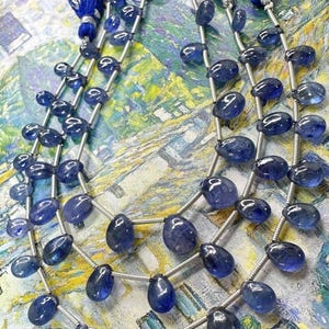 Può includere: Più fili di perline di pietre preziose blu con filo color argento, ogni filo termina con un nappa di fili blu. Le perline sono ovali e a forma di goccia, con finitura lucida. Lo sfondo è un motivo astratto colorato.