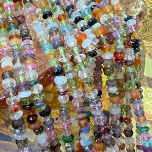 May include: Close-up of multiple strands of colourful gemstone beads. The beads are in a variety of shapes and sizes, with a mix of pink, green, orange, and blue hues. The beads have a faceted or polished finish and are strung together.