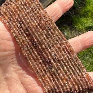 May include: A collection of small, brown, faceted gemstones strung together. The beads display a range of brown hues, creating a shimmering effect. The gemstones are held in a hand, with a blurred background of green grass and wooden planks.