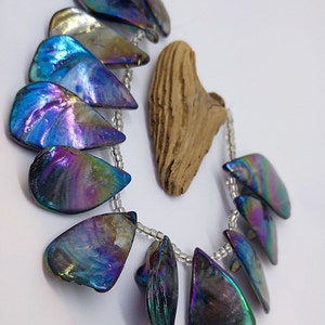 Puede incluir: Un collar hecho de cuentas iridiscentes de color azul y morado en forma de lágrima enhebradas en una cadena plateada. Las cuentas tienen un acabado nacarado y están dispuestas en un patrón escalonado.