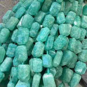 May include: Strands of Amazonite gemstone beads. The beads are a light turquoise colour, with a faceted, rectangular shape. The beads are strung on a green cord. The gemstones are approximately 1.5 cm in length.