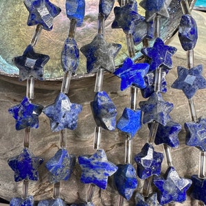 Op de afbeelding: Detailopname van lapis lazuli kralen. De kralen zijn stervormig en langwerpig, in diepe blauwtinten met grijze marmering. Ze zijn geregen op transparante koorden. De achtergrond is neutraal grijs.