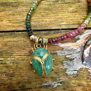 May include: A green gemstone beetle pendant with gold accents hangs from a multi-colored beaded necklace. The necklace features pink, green, blue, and black beads.