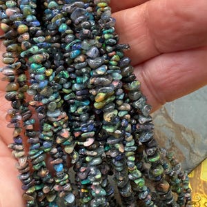 May include: Close-up of multiple strands of small, polished gemstones. The gemstones are in shades of blue, green, pink, and black. They are strung together, creating a textured, iridescent effect. These are likely for jewellery making.