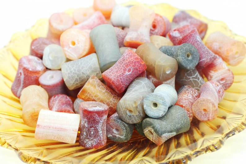 May include: A collection of agate beads in various shades of red, orange, and grey. The beads are all cylindrical and have a rough, textured surface.