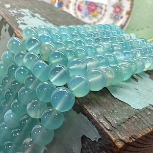 Könnte beinhalten: Eine Strähne aus hellblauen runden Edelsteinperlen. Die Perlen sind glatt und durchscheinend, mit einem leichten Schimmer. Die Perlen sind lose auf einer Holzoberfläche angeordnet.