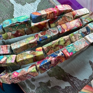 May include: Strands of rectangular gemstone beads in shades of green, blue, pink, and brown. The beads have a natural, textured appearance, and are strung together. The beads are approximately 2.5 cm long.