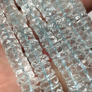May include: Close-up of several strands of faceted, translucent aquamarine beads. The beads are strung together with a light blue thread, creating a shimmering effect. The beads have a geometric, multi-faceted design.