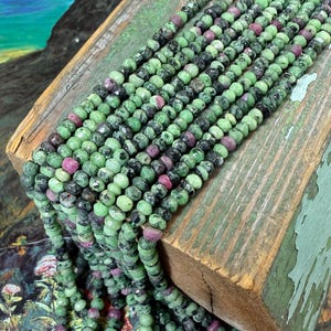 May include: Strands of gemstone beads in shades of green, black, and pink. The beads are small and round, and they are strung together to create a necklace or bracelet. The beads are displayed on a weathered wooden surface.