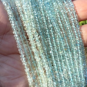 May include: Multiple strands of aquamarine gemstone beads in shades of blue and clear. The small, round beads are strung together, creating a delicate, shimmering effect. The beads are held in a hand, with a blurred background.