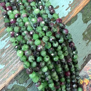 Puede incluir: Primer plano de hilos de cuentas de piedras preciosas en verde, negro y burdeos. Las cuentas redondas y facetadas están ensartadas. Estas piedras preciosas probablemente se utilizan para la fabricación de joyas.