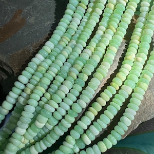 Könnte beinhalten: Eine Strähne aus hellgrünen Edelsteinperlen, jede Perle ist oval geformt und hat eine glatte, polierte Oberfläche. Die Perlen sind in einem lockeren Haufen angeordnet, der die Variation in Farbe und Textur zeigt.