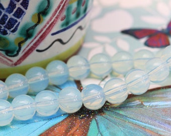 May include: A strand of pale blue opal beads, each bead is round and translucent. The beads are arranged in a circular pattern, with some beads overlapping. The beads are on a surface with a blue and green pattern.