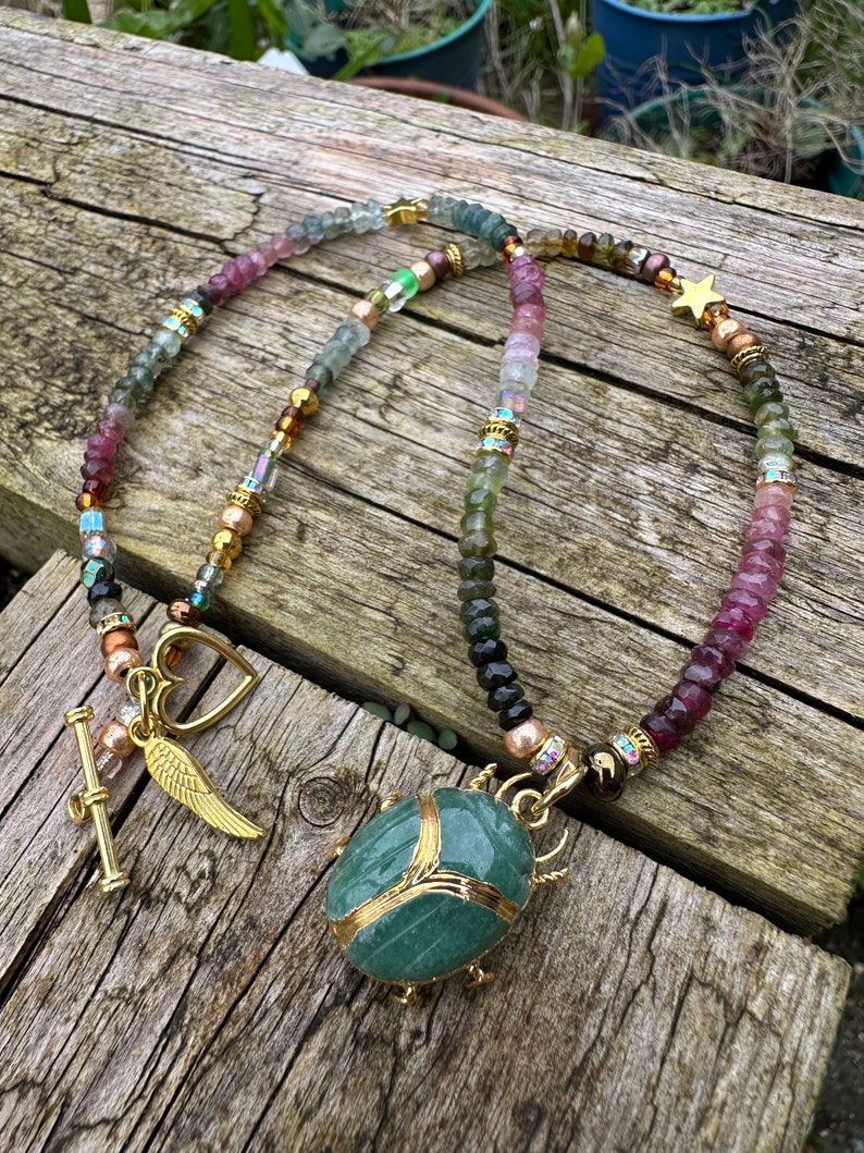 May include: A multi-colored beaded necklace with a green stone beetle pendant, gold accents, and charms including a heart, wings, and a star.