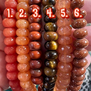 Puede incluir: Seis pulseras diferentes hechas de piedras naturales. Las pulseras están dispuestas en fila, siendo la primera roja, la segunda naranja, la tercera roja, la cuarta marrón y dorada, la quinta rosa y la sexta marrón. Cada pulsera tiene un número del 1 al 6.