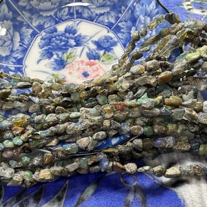 May include: Strands of rough-cut gemstone beads in shades of green, brown, and grey. The beads are displayed on a blue and white floral patterned plate. The beads are likely for jewellery making.