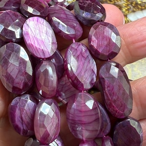 May include: A string of faceted, oval gemstones in shades of deep pink and purple. The stones have a polished, reflective surface, and are strung together, suggesting a necklace or bracelet.