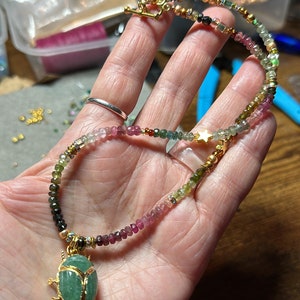 May include: A multi-colored gemstone necklace with a gold beetle pendant. The necklace features a variety of green, pink, and black gemstones, as well as gold beads and a gold star charm.