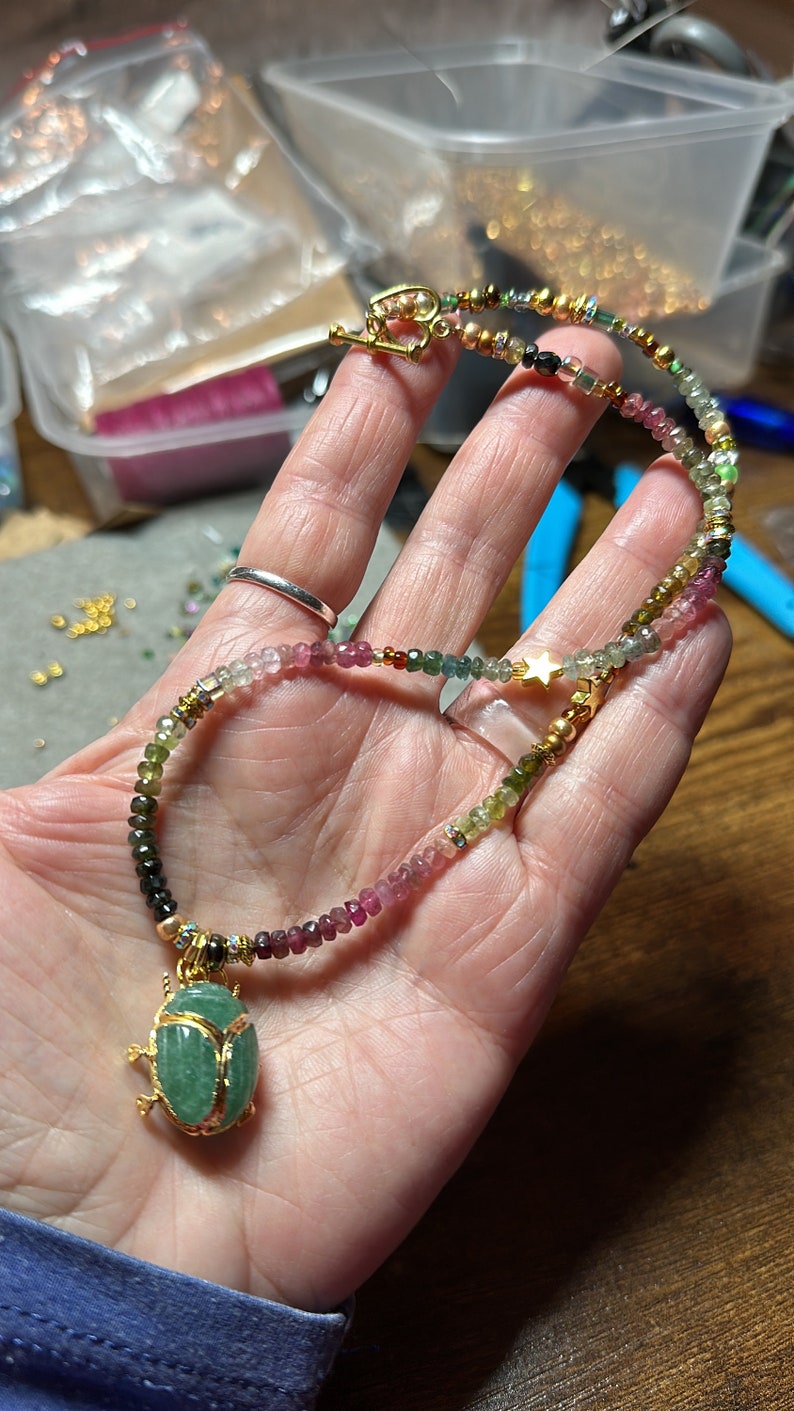 May include: A multi-colored gemstone necklace with a gold beetle pendant. The necklace features a variety of green, pink, and black gemstones, as well as gold beads and a gold star charm.