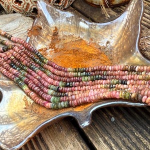 Puede incluir: Cadenas de cuentas de piedras preciosas multicolores, principalmente rosas, verdes y marrones, se muestran en un plato en forma de hoja. Las cuentas son pequeñas y redondas, lo que sugiere un collar o una pulsera. El plato tiene un brillo metálico.