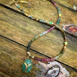 May include: A gold necklace with a green gemstone beetle pendant. The necklace is made of various colored beads, including pink, green, and brown. The beads are interspersed with gold stars.