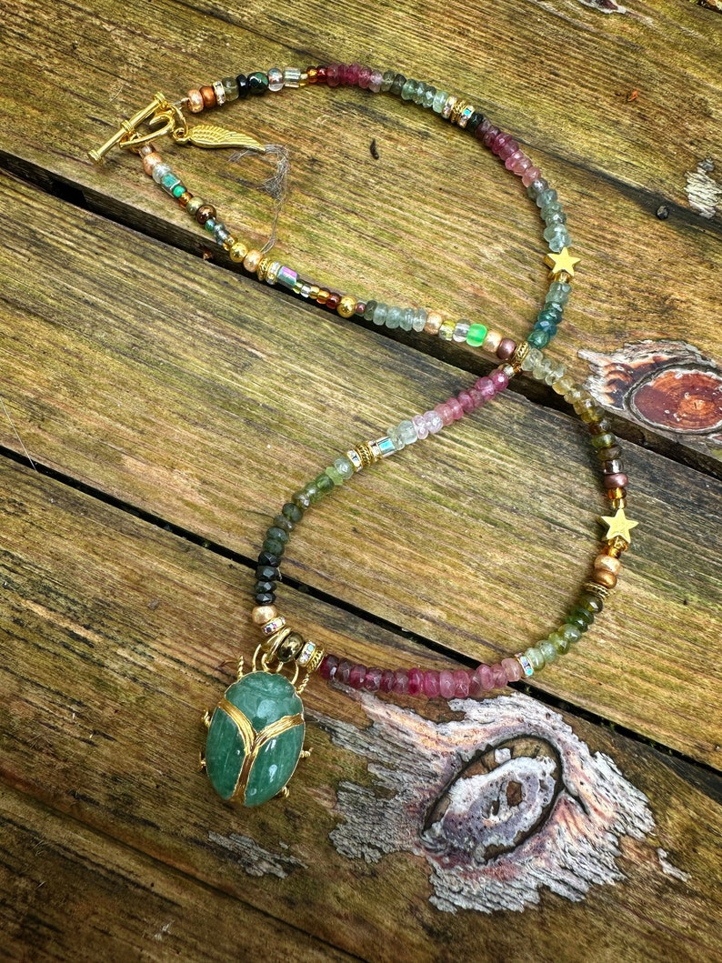 May include: A gold necklace with a green gemstone beetle pendant. The necklace is made of various colored beads, including pink, green, and brown. The beads are interspersed with gold stars.