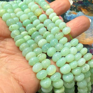 May include: A collection of light green gemstone beads. The beads are oval and polished, strung on a green thread. The beads' colour is a soft, pastel green. The beads are held in a hand. Each bead is approximately 8mm in diameter.