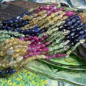 May include: A collection of gemstone bead necklaces in various colors, including blue, green, yellow, and pink. The beads are oval-shaped and strung together. The necklaces are displayed on a dark gray stone and a green glass dish.