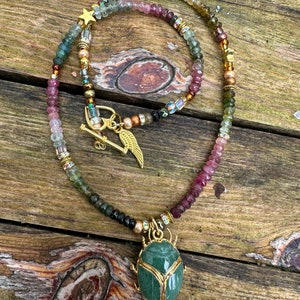 May include: A gold necklace with a green stone beetle pendant. The necklace is made of multi-colored beads, including pink, green, and brown.