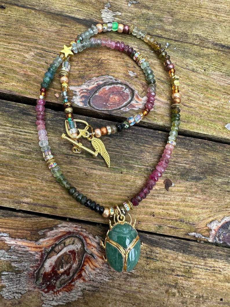 May include: A gold necklace with a green stone beetle pendant. The necklace is made of multi-colored beads, including pink, green, and brown.