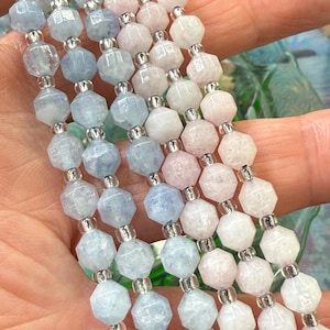May include: Close-up of multiple strands of gemstone bead bracelets. The beads are faceted and in shades of light blue, pink, and white. Silver-coloured metal beads separate the gemstone beads. The bracelets are held in a hand.