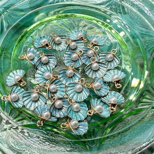May include: A collection of teal and gold seashell charms with faux pearl centers, arranged in a clear, green glass dish. The charms have a fan-like design and are suitable for jewelry making or craft projects.
