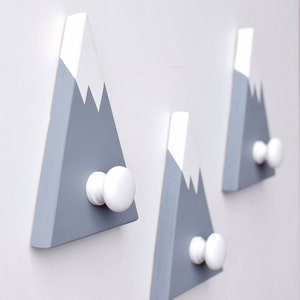 May include: Three gray and white wooden mountain-shaped hooks with white knobs. The hooks are mounted on a white wall.