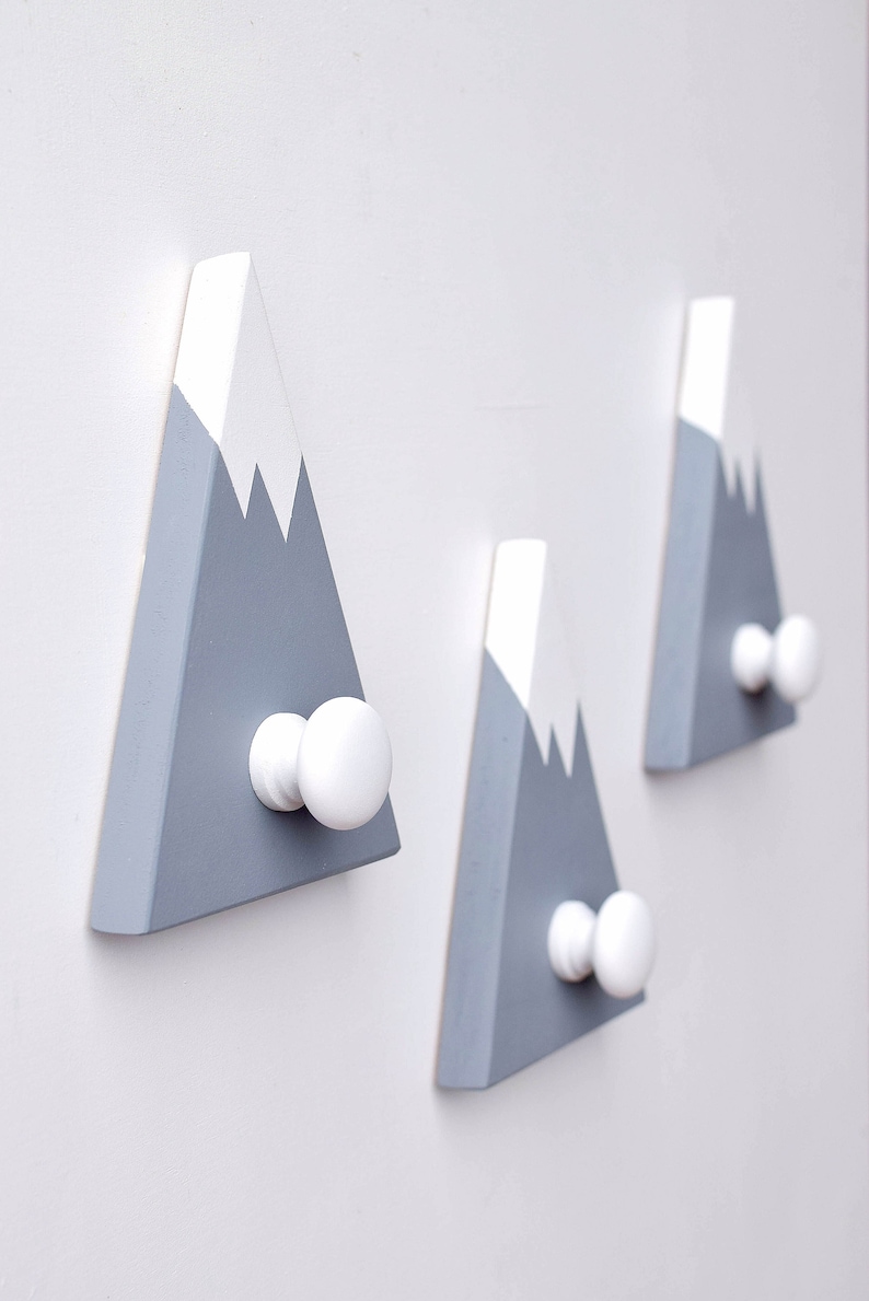 May include: Three gray and white wooden mountain-shaped hooks with white knobs. The hooks are mounted on a white wall.
