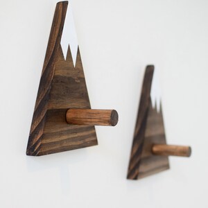 May include: Two wooden mountain-shaped hooks with a white snowcap on top. The hooks are mounted on a white wall.