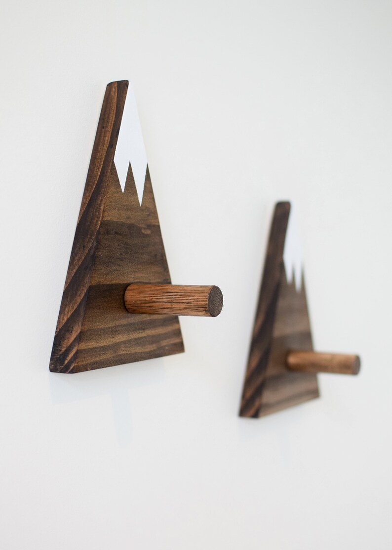May include: Two wooden mountain-shaped hooks with a white snowcap on top. The hooks are mounted on a white wall.