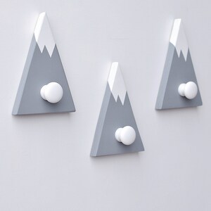 May include: Three gray wooden mountain-shaped hooks with white snow caps and white knobs. The hooks are mounted on a white wall.